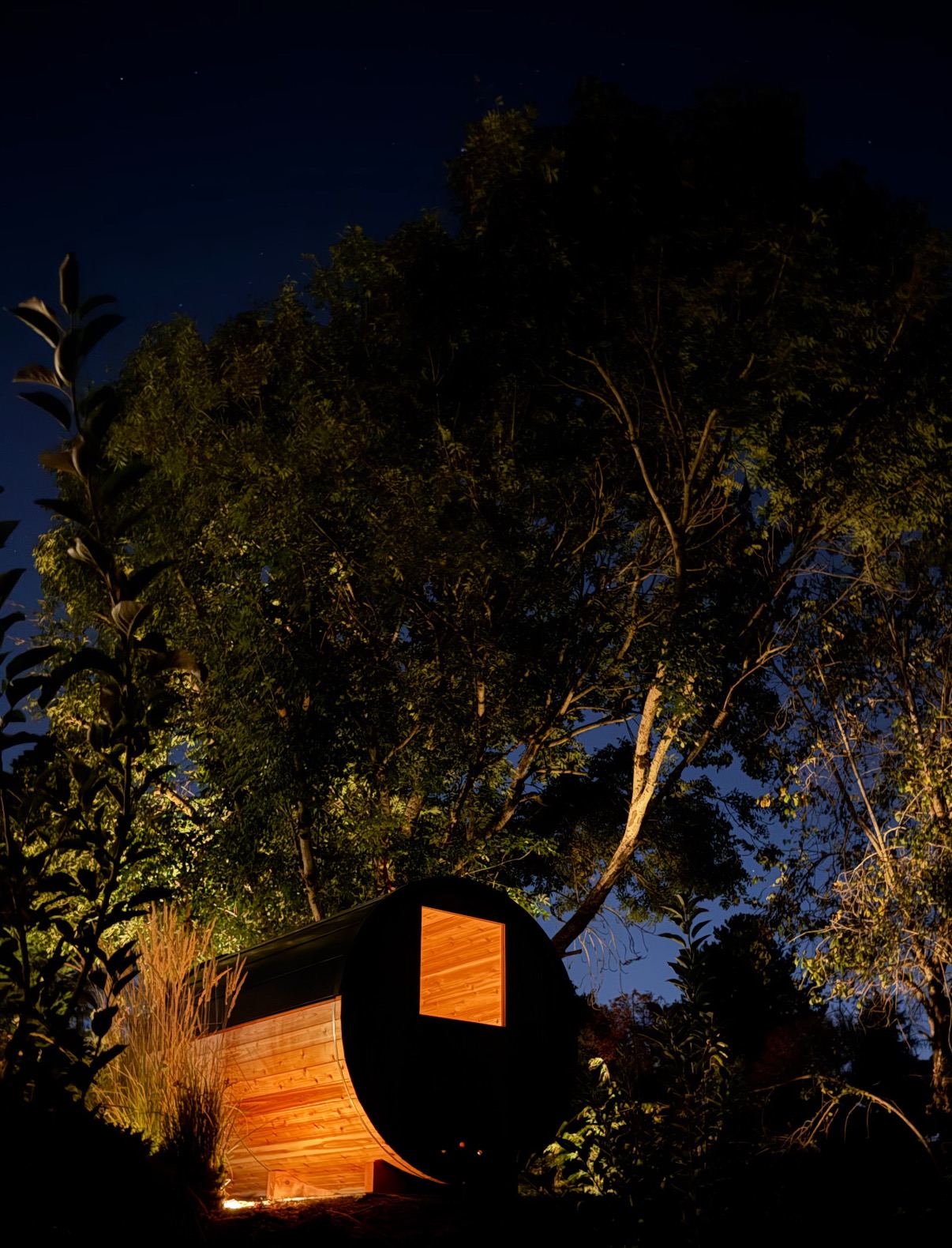 The cedar barrel sauna glowing under tall trees at night