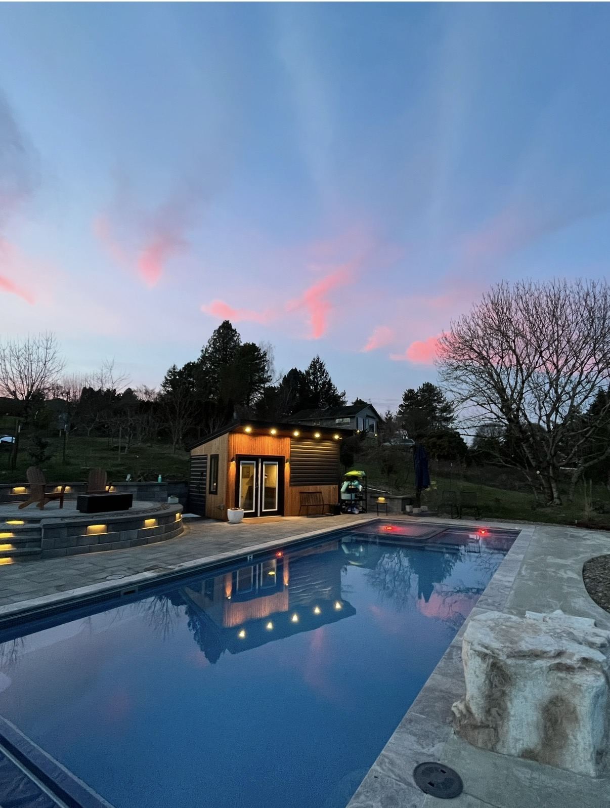 The lit cedar pool house reflected in the pool under a pink dusk sky